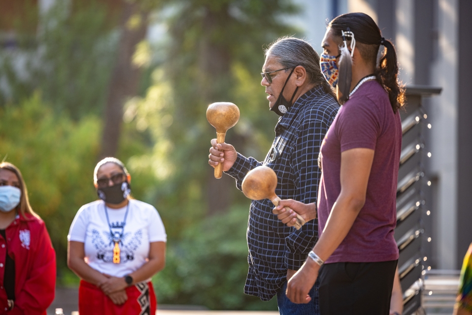 Two Native American men shake gourd instruments while performing a semester blessing ceremony