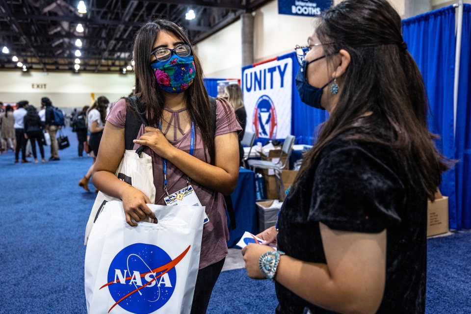 Two young women speak at astronomy conference with booths behind them and one woman holding a NASA bag