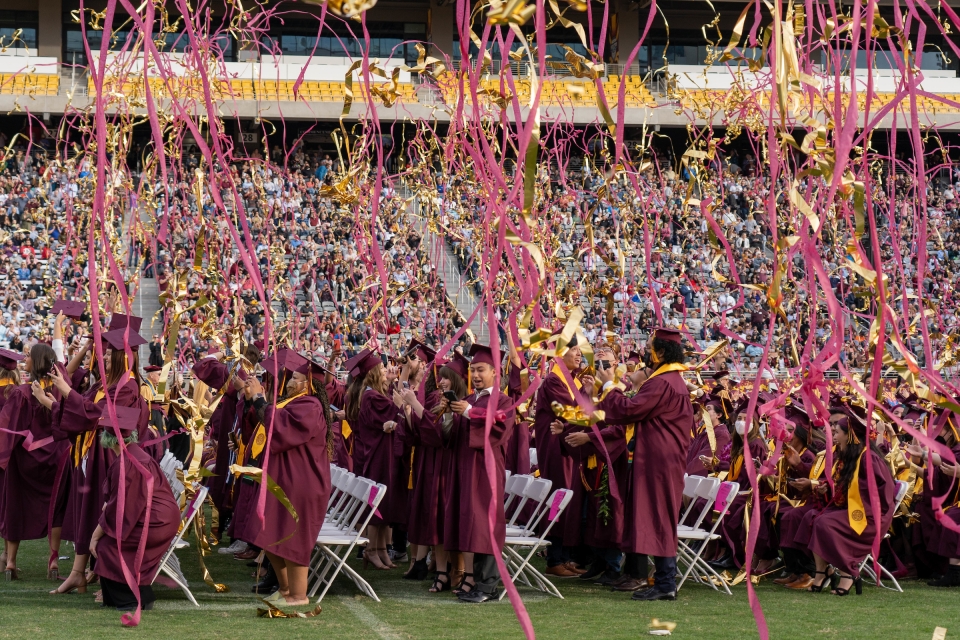 Maroon and gold streamers rain down on a crowd of students in graduation gowns on a football field