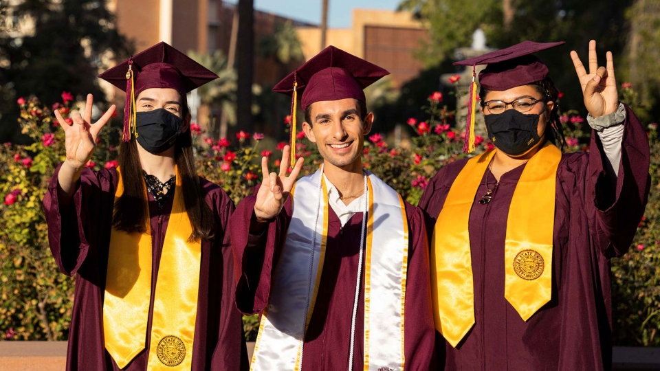 Three students in graduation gowns and caps make the pitchfork gesture; one is unmasked and two have masks on