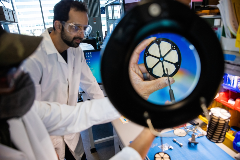 Two men in a lab point at a circular air filter framed by a magnifying lens