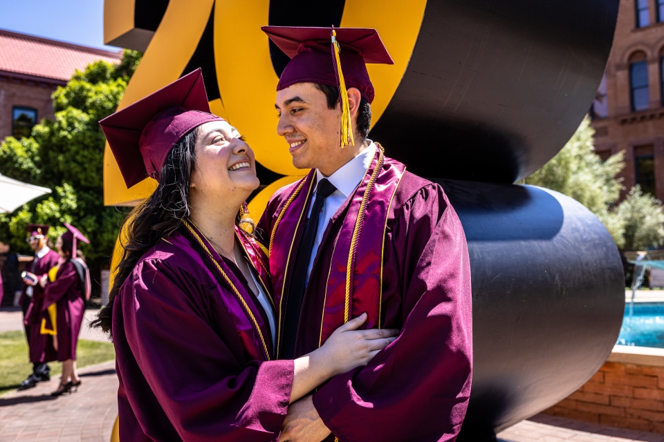 A couple in graduation caps and gowns look at each other and smile while posing in front of a giant 2022 sign