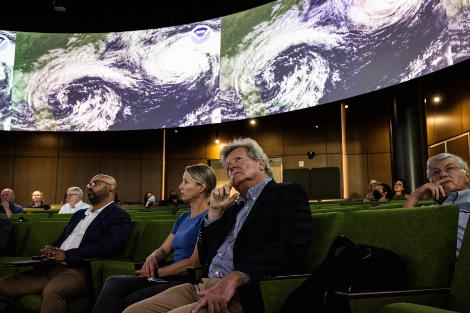 People in audience chairs look up at screens showing satellite footage of clouds over oceans