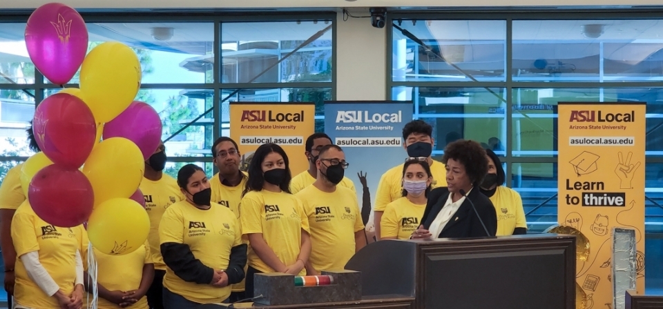 Woman speaking into a microphone while a group of people wearing gold ASU Local shirts look on.