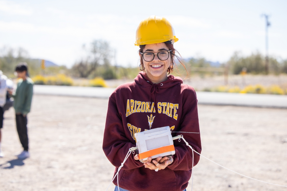 Student wearing hard hat and ASU sweatshirt holds a weather balloon payload