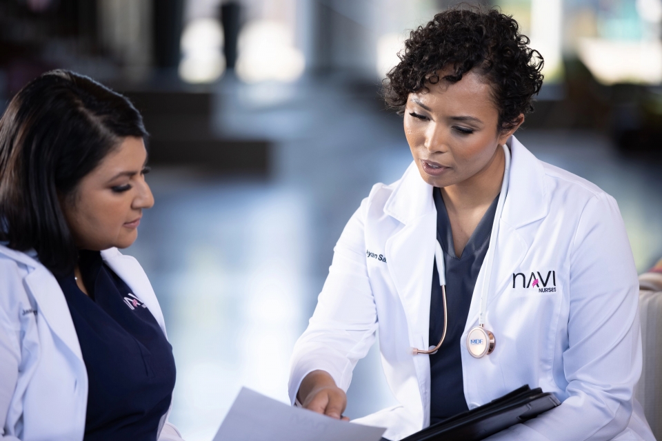 Ayan Said wears a white coat with NAVI on it and a stethoscope around her neck. She points to a piece of paper that her colleague Jasmine Bhatti is looking at. Bhatti is also wearing a white coat with NAVI on it and navy scrubs
