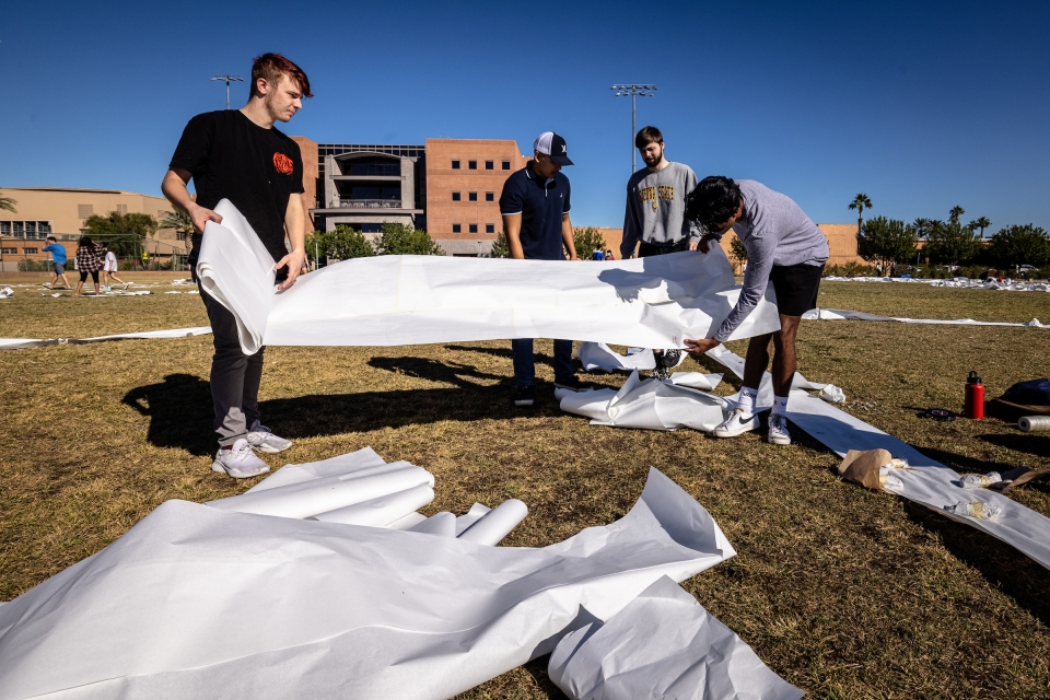 ASU architecture students unrolling a large roll of butcher paper on a grassy field.
