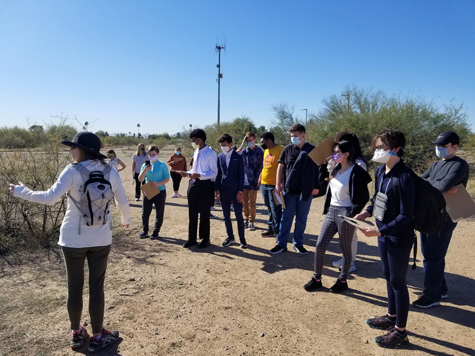 Group of students gathered in a desert setting, listening to an instructor.