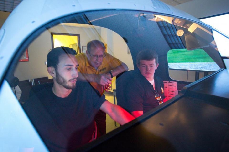 two men seated in training cockpit as instructor looks on