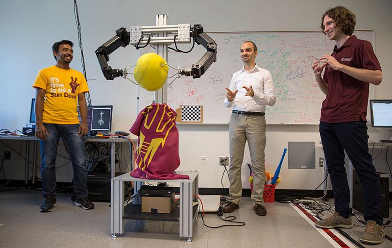 Three men in a lab crowd around a robot clutching a ball. The caption reads: "I’m always having a blast with my students, coming up with crazy ideas,” says Heni Ben Amor (center), shown here with Yash Rathore (left) and Kevin Luck.