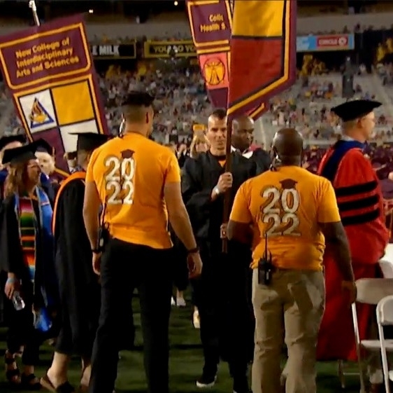 Marcus Jones wears a gold shirt that reads "2022" on the back as he directs people carrying gonfalons on a football field during a graduation ceremony.