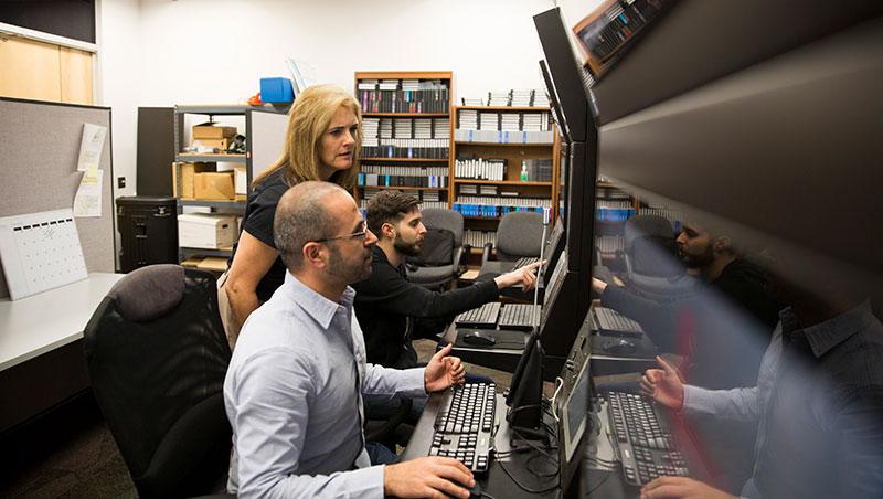 Three people, in a lab, peer at a row of monitors. The caption reads: Nancy Cooke works with Aaron Bradbury (left) and Mustafa Demir on the Synthetic Teammate project. Photo courtesy of the Ira A. Fulton Schools of Engineering.