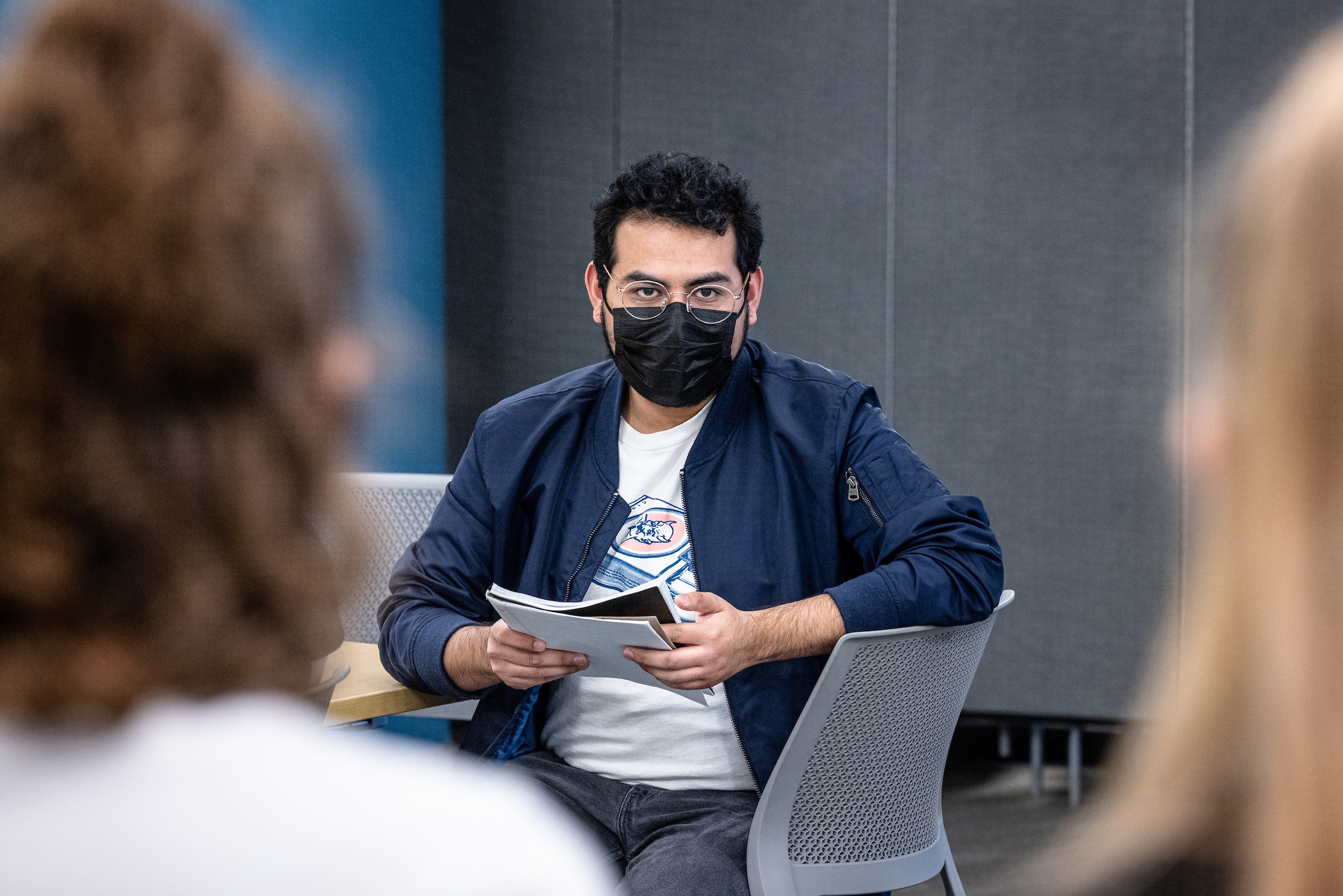 man sitting in a chair wearing a face mask and reading from a notebook