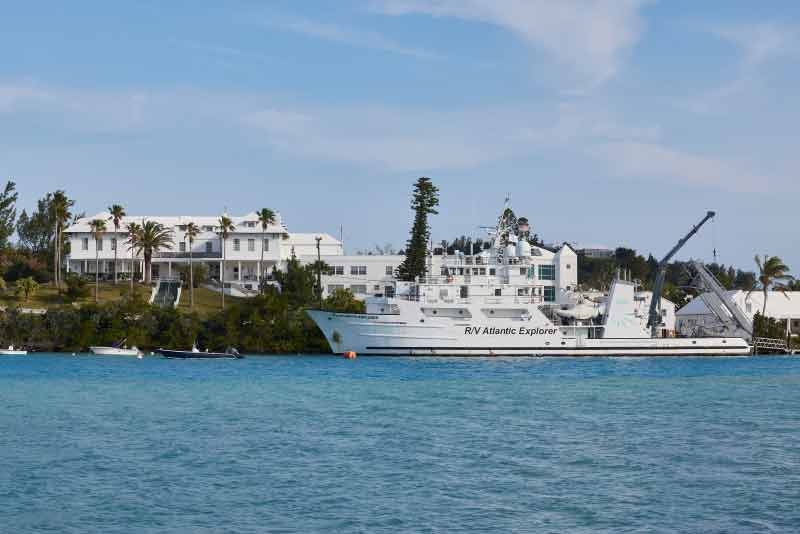 A large white boat docked next to an island with smaller boats and a large building nearby.