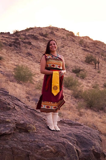 Woman wearing traditional Native American dress, standing on a rock at the base of a butte.