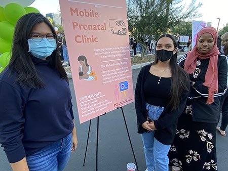 Edson College students stand next to their poster during the Health Innovation Exhibition. The poster is a pale pink and has the words Mobile Prenatal Clinic on it