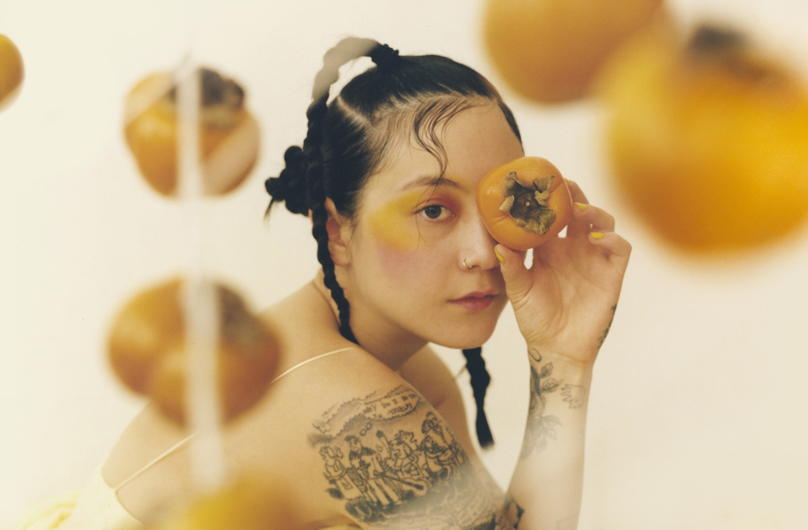 Michelle Zauner looks over her shoulder, covering one eye with a golden persimmon. She wears golden eye makeup and styled hair braids. Persimmons float around her out of focus.