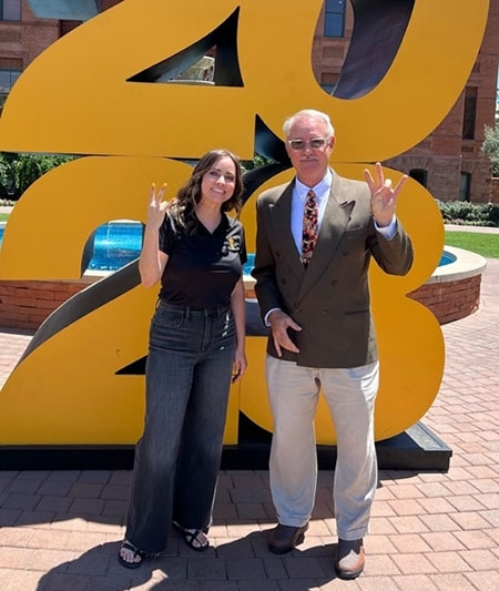 A woman and a man standing in front of a giant 2023 sign make the ASU pitchfork hand gesture