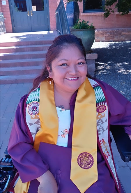 A woman wearing a graduation gown and a big smile sits in a wheelchair outside a building