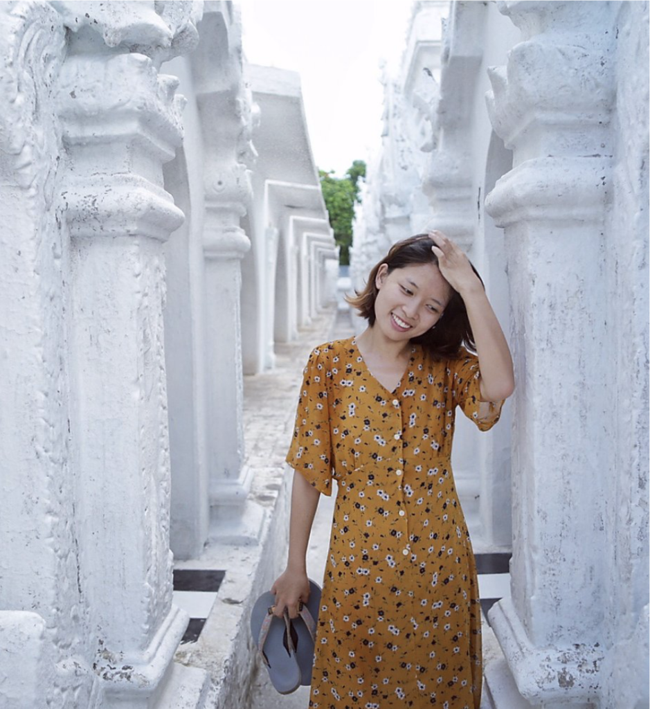 Beiyin Deng with white marble behind her. She is holding a pair of flip-flop shoes and is wearing a yellow dress.