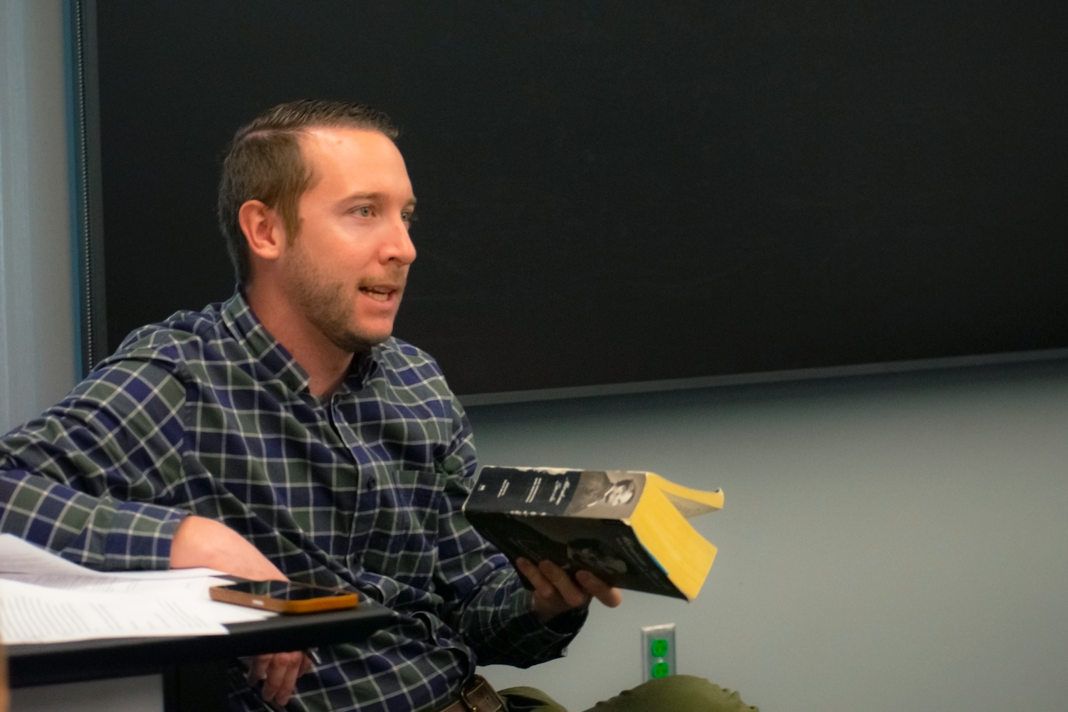 ASU Assistant Professor Zachary German seated at the front of a class, speaking while holding a book.
