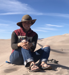 ASU PhD student Madeline Kelley wearing a hat, jeans and sandals while sitting in a sandy expanse.