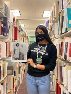 Person wearing a mask and holding a photograph while standing amongst bookshelves