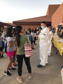 Eli Fox, wearing a white jumpsuit and blue surgical face mask, speaks with a group of elementary school students who are facing away from the camera. The group is gathered outside a school building.