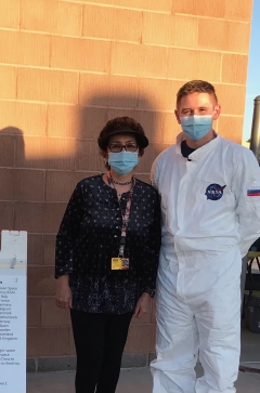 Saule Moldabekova Robb, left, and Eli Fox stand in front of a brick wall. She is wearing a dark blouse and pants and has dark hair and is wearing glasses and a blue face mask. He is wearing a mask and a white jumpsuit with a NASA logo on the breast/