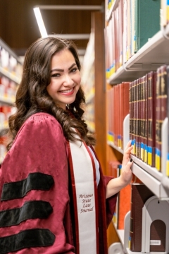 ASU Law alum Claire Newfeld wearing a graduation gown and standing next to a stack of books in a library.