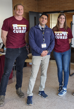 Umar Suleyman stands in khaki pants and a navy pullover. He is looking at the camera with his hands folded in front of his body. A man and a woman stand on either side of him wearing blue jeans and maroon shirts that say "Monolingualism can be cured."