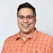 ASU directory photo of new faculty member Derek Pacheco, who smiles at the camera while wearing a checked orange shirt and stands in front of a light grey background.