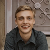 Portrait of a smiling young person in a dark grey shirt with a decorative wall in the background.