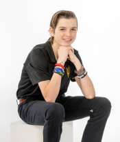 Young person sitting on a white cube, wearing a black polo shirt with a pitchfork emblem and dark jeans, smiling at the camera.