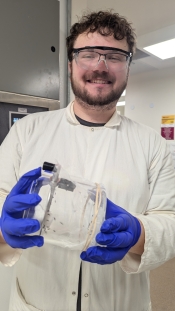 A man in a lab coat, blue gloves, and safety goggles holds up a container full of mosquitoes while smiling at the camera.