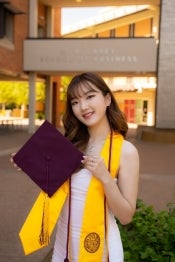 Young woman posing for a graduation photo in front of the ASU school of business holding a maroon graduation cap and wearing a gold stole