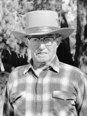 Black and white photo of an older man in a hat wearing glasses and a plaid button up shirt