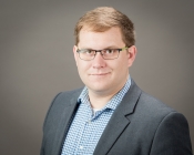 ASU Assistant Professor Aaron Guest smiles at the camera for a professional portrait. He is wearing a checkered blue and white button up shirt and a gray blazer.