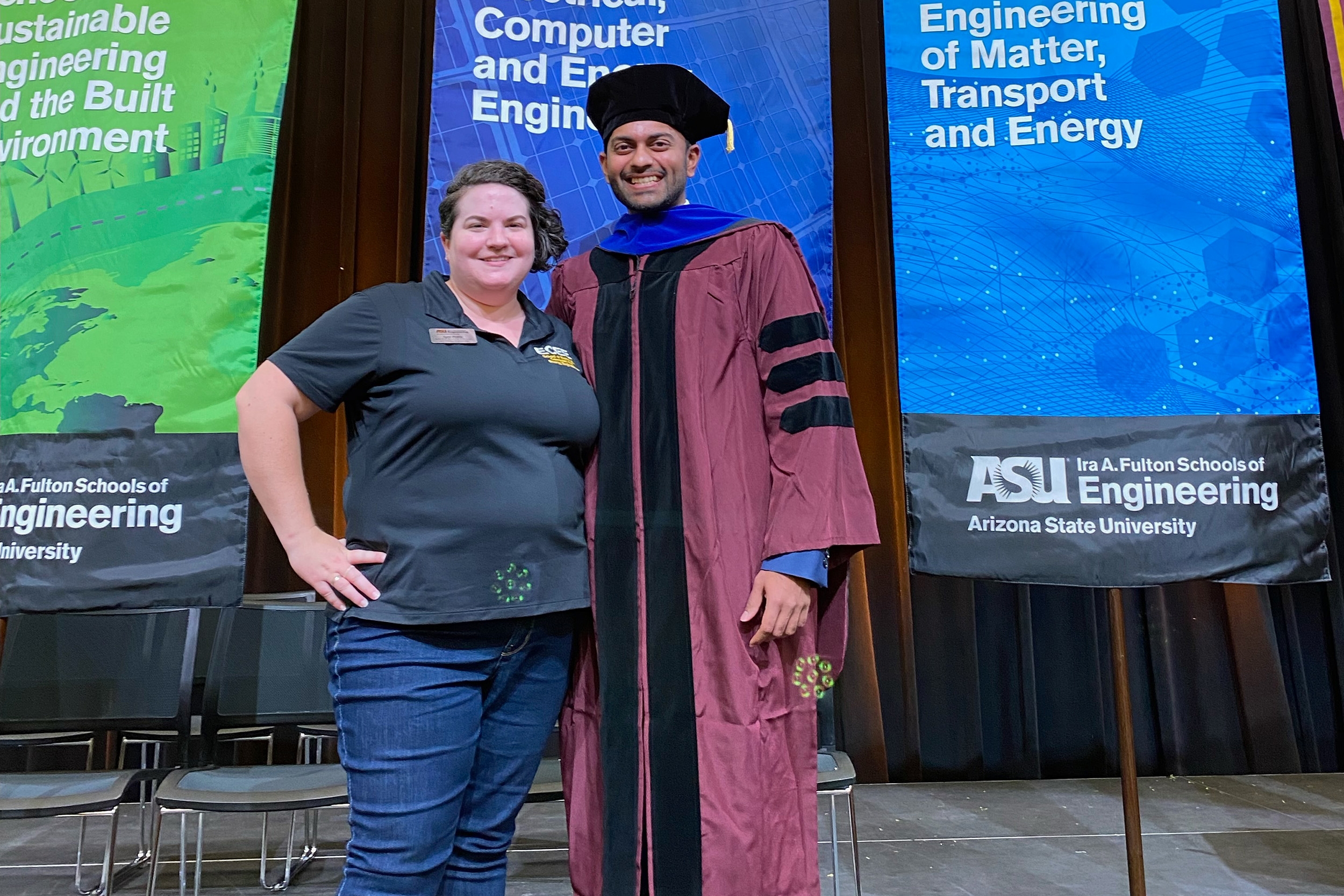 Lynn Pratte poses to the left with Venkatraman Balasubramanian, who is wearing a graduation gown at stole, on stage with ASU banners behind them.