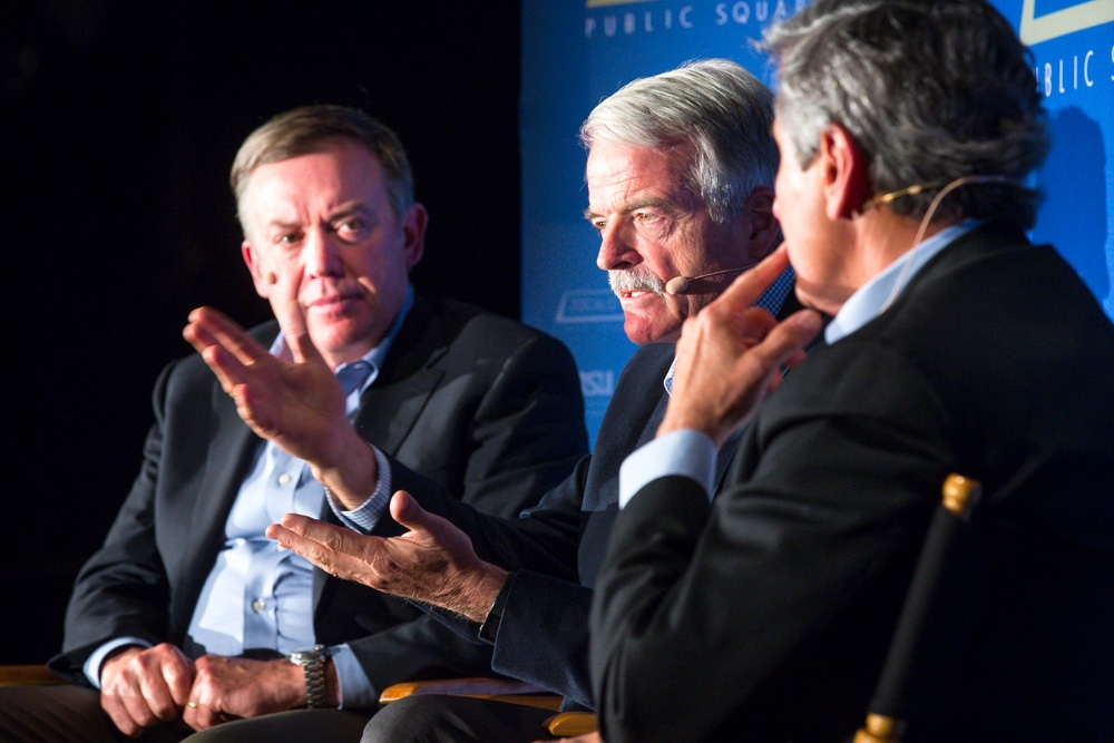 Three men discuss universal health care on a stage.