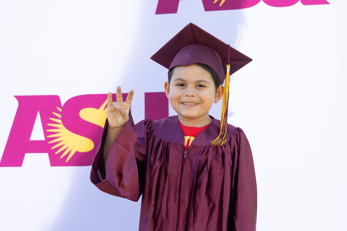 A young boy wearing a graduation cap and gown holds up the ASU pitchfork gesture in front of an ASU backdrop