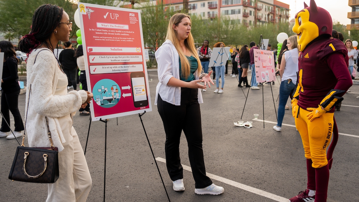 ASU students stand next to a research poster while talking to Sparky the Sun Devil mascot.