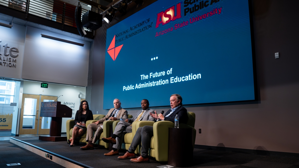 One woman and three men seated on a stage with a screen behind them 