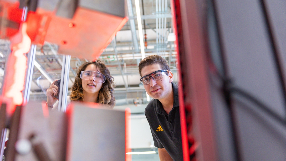 Two students look at a gray robotics machine that is glowing red
