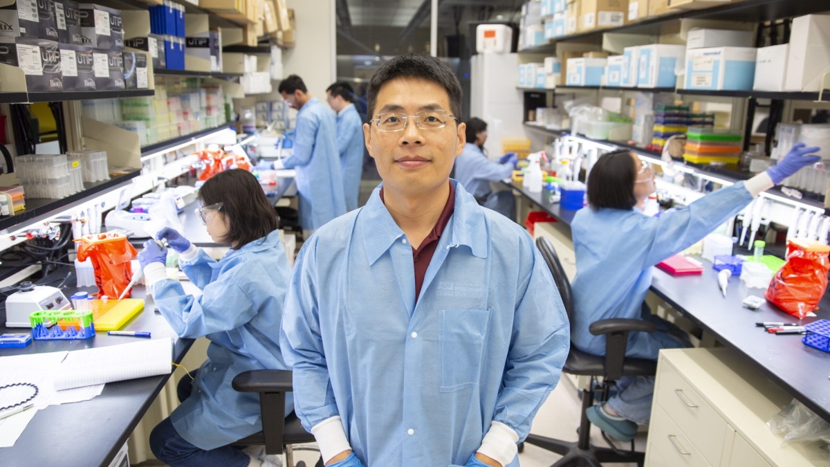 A man wearing glasses and a blue lab coat poses for a photo in a lab with researchers working behind him