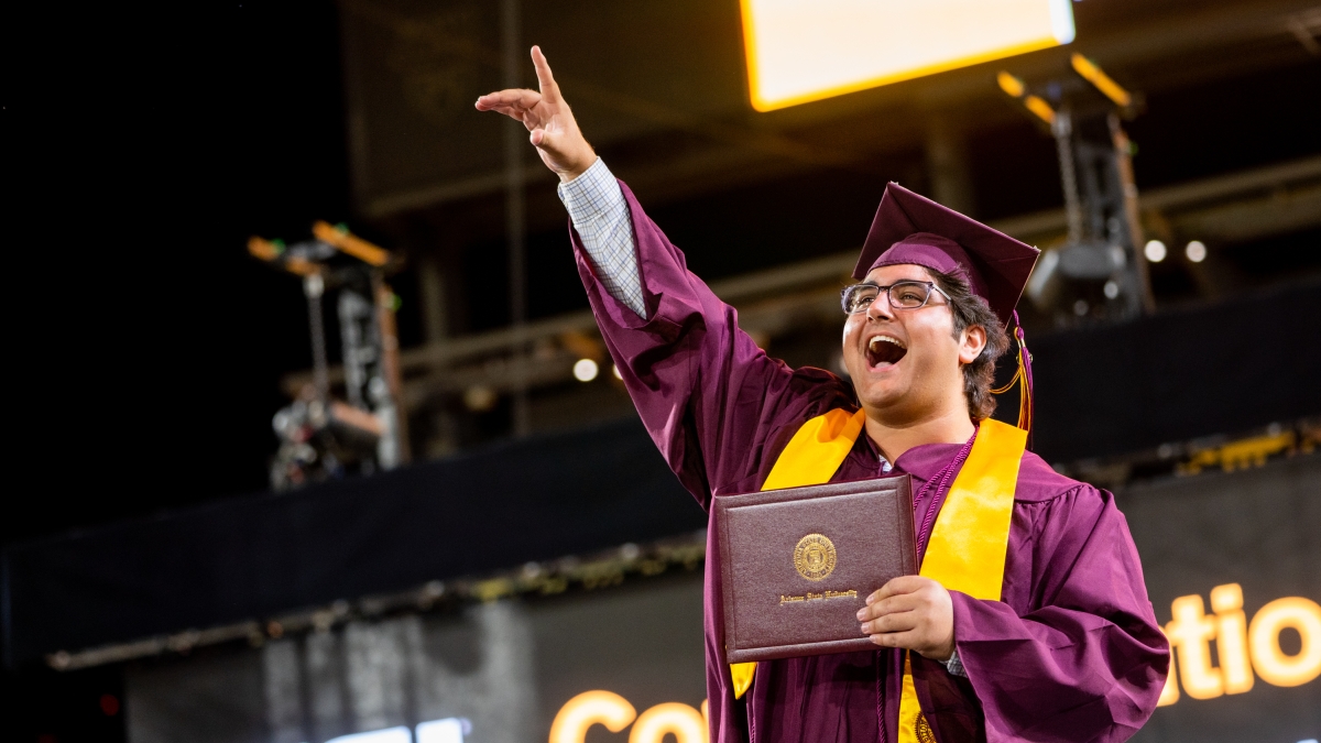 An ASU graduate in cap and gown cheers while holding his diploma at graduation