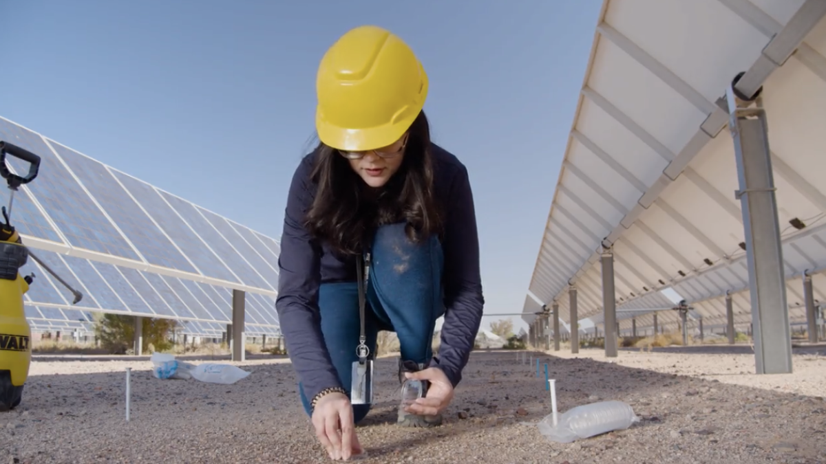 A woman in a yellow construction hat kneels next to solar panels.