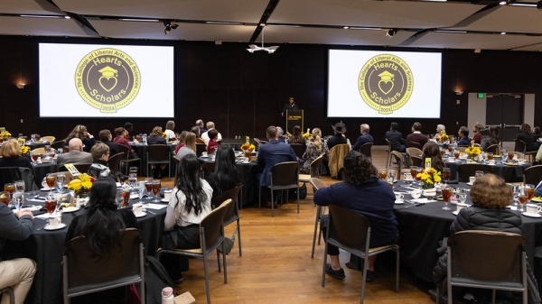 People seated in an event space where a man is speaking on a stage next to screens that display the words "Hearts Scholars."