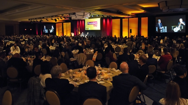 Audience seated at round tables in a large room with a stage and a screen that reads "Founders' Day."