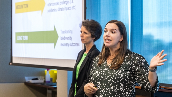Two women stand in front of a screen talking to a crowd